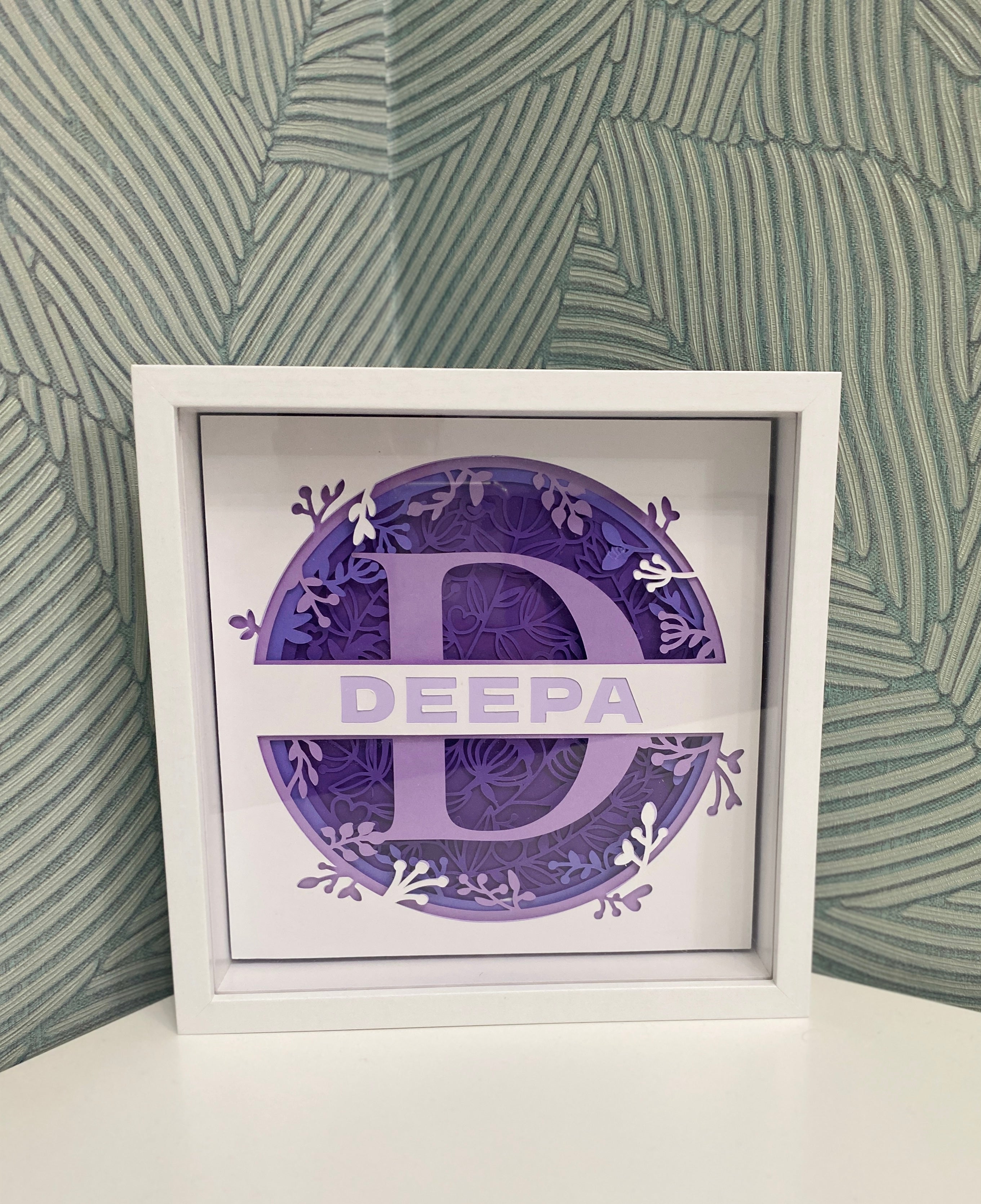 Framed decorative letter 'D' with 'DEEPA' text against a patterned wall.